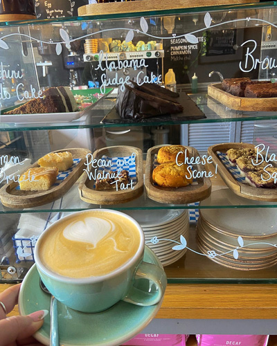 An image of someone holding up a latte in front of a cake cabinet containing various gluten free cakes at Emzi, a cafe in Clifton, Bristol
