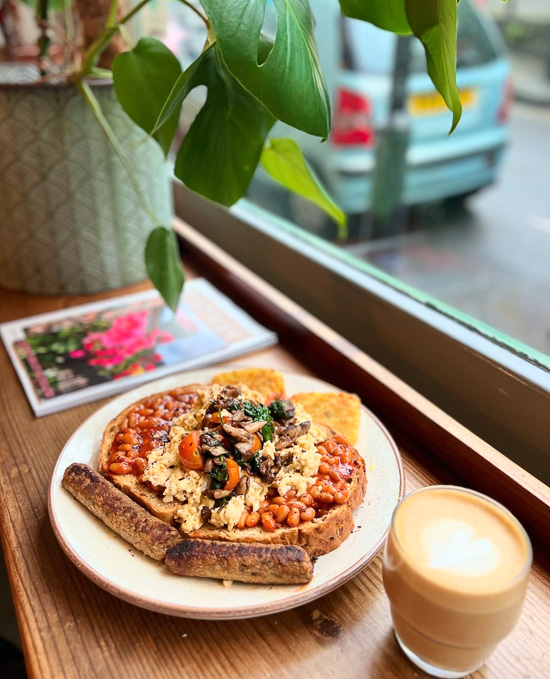 An image showing a plate of food containing toast, scrambled eggs, baked beans, hash browns, mushrooms and tomatoes as well as coffee, on a windowsill at Emzi.